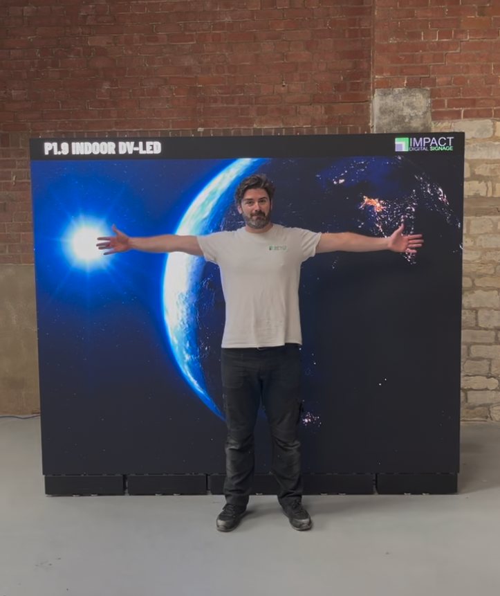 A man stands with his arms outstretched in front of a large black display screen showing a blue Earth and space scene. The screen has text reading 'P1.9 Indoor DV-LED 