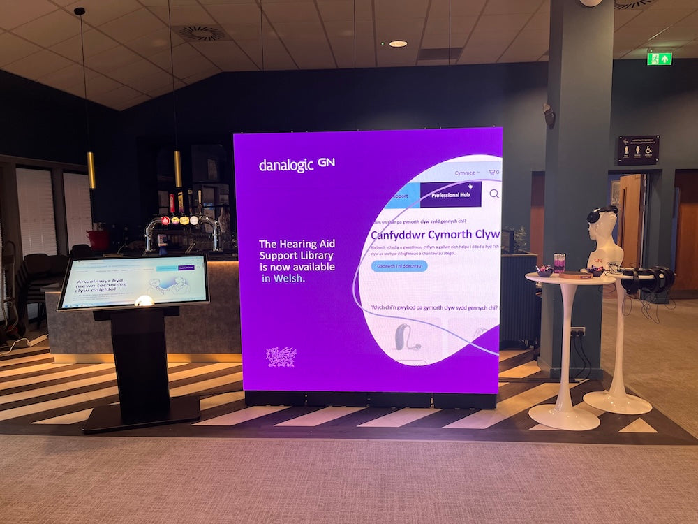 A large digital display screen in a modern indoor lobby shows a purple and white interface with text about hearing aids. The room has dark walls, a patterned floor, a smaller side screen on a pedestal, and a white mannequin head on a table.