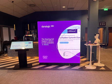 A large digital display screen in a modern indoor lobby shows a purple and white interface with text about hearing aids. The room has dark walls, a patterned floor, a smaller side screen on a pedestal, and a white mannequin head on a table.