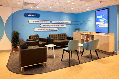 Modern office waiting area with blue walls, couches, chairs, and a digital display.
