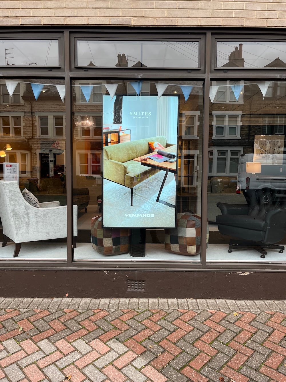 A large vertical poster for 'Smiths of Harrogate' is displayed on the window of a furniture store, showing a light green sofa and a patterned rug. The storefront has large dark-framed windows reflecting the street and other furniture inside.