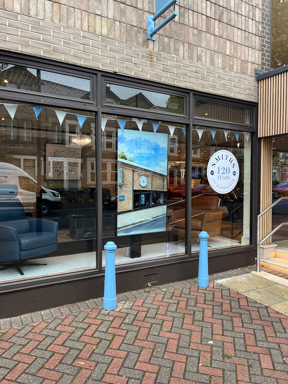 A brick building storefront with large black-framed windows, blue bollards on the sidewalk, and a round white sign reading 'SOUTH 120'. The window displays a blue interior scene and bunting, with reflections of the street and parked vehicles.