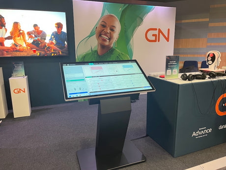 A modern touch kiosk display stand with a large angled screen showing a software interface, set in a corporate exhibition booth with GN and Advance branding.